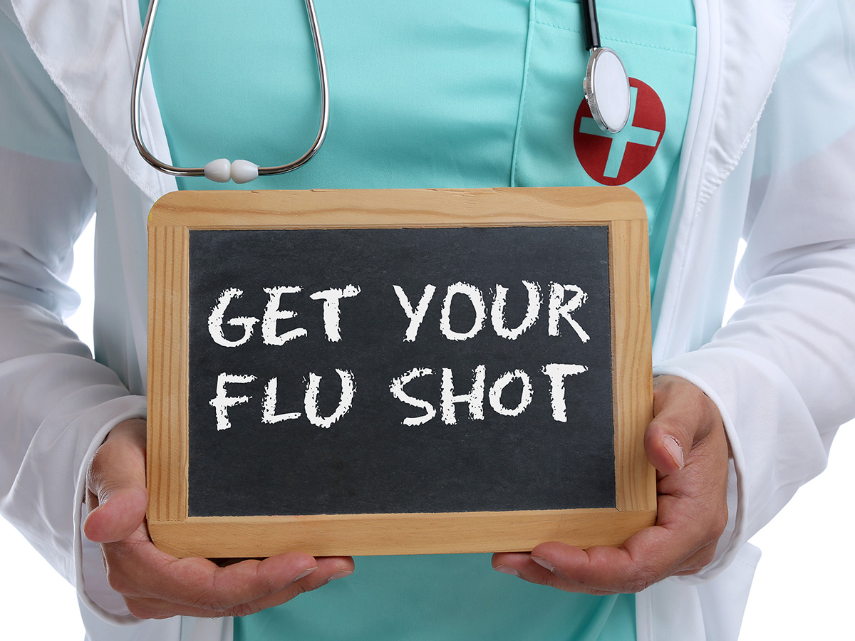 Female doctor with white coat and stethoscope holding a chalkboard sign that reads “GET YOUR FLU SHOT”